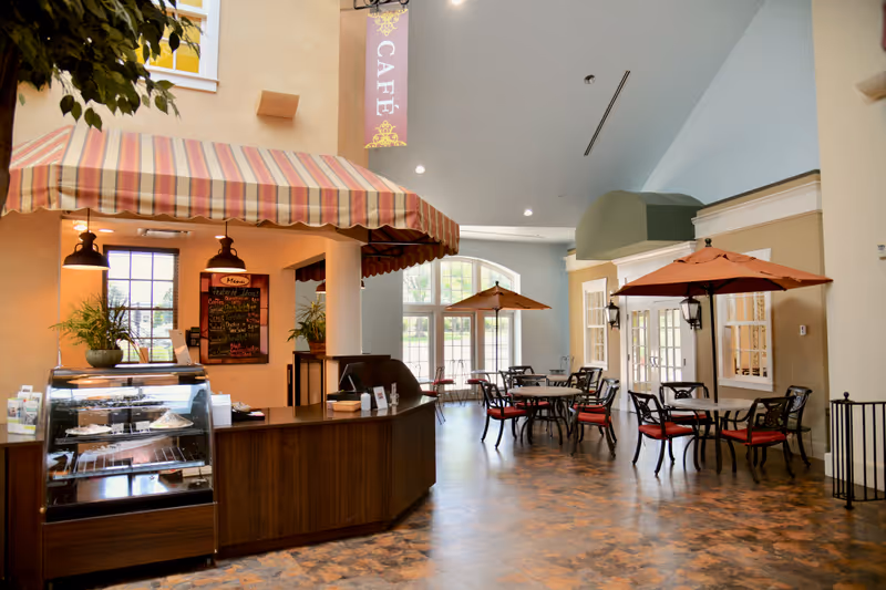 Interior view of a café area in a senior living facility with a striped awning over the counter, a display case with pastries, a menu board, and several tables with chairs and umbrellas. Large windows allow natural light to fill the space.