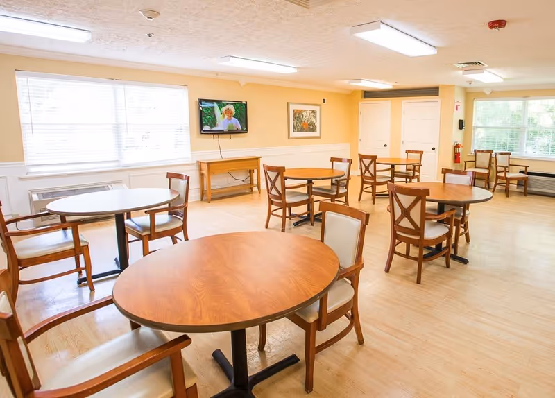 A bright dining room with several round wooden tables and chairs arranged neatly. The room has large windows with blinds, light-colored walls, and a wall-mounted television displaying a person. There is a framed picture on the wall and a fire extinguisher near a set of double doors.