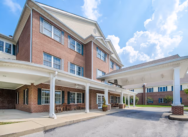 Brick multi-story senior living building front with a covered porte-cochere, outdoor seating, and clear blue sky.