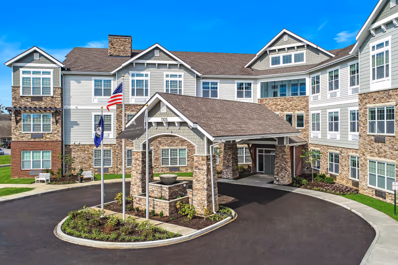 Exterior view of a senior living facility named The Ashton on Dorsey, featuring a large covered entrance with stone pillars, multiple windows, and three flagpoles with flags in front of the building under a clear blue sky.
