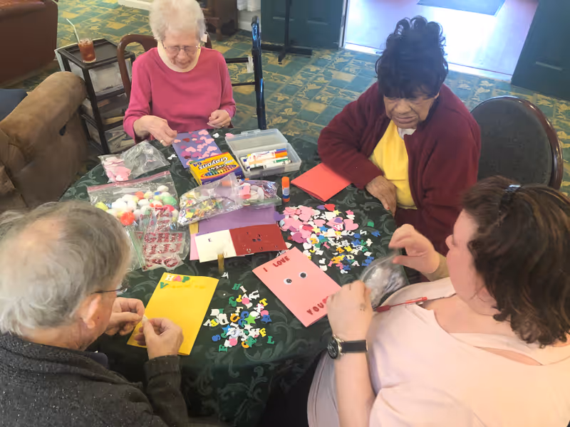Four elderly individuals sitting around a table engaged in a craft activity, making colorful cards with letters, pom-poms, and other decorative materials in a cozy room with patterned carpet.