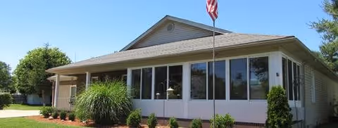 Front exterior of a single-story assisted living facility with large windows, manicured landscaping, and an American flag on a flagpole.