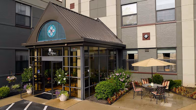 Entrance to a building with a glass vestibule featuring a decorative stained glass window above the door. There are potted plants and hanging flower baskets around the entrance. To the right, there is an outdoor seating area with a round table, four chairs, and a large beige umbrella. The building exterior is painted in neutral tones with windows visible.