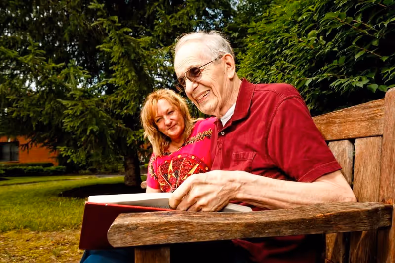 An elderly man and a middle-aged woman sitting on a wooden bench outdoors, with the man holding and looking at a large book while the woman smiles beside him. They are surrounded by green trees and grass.