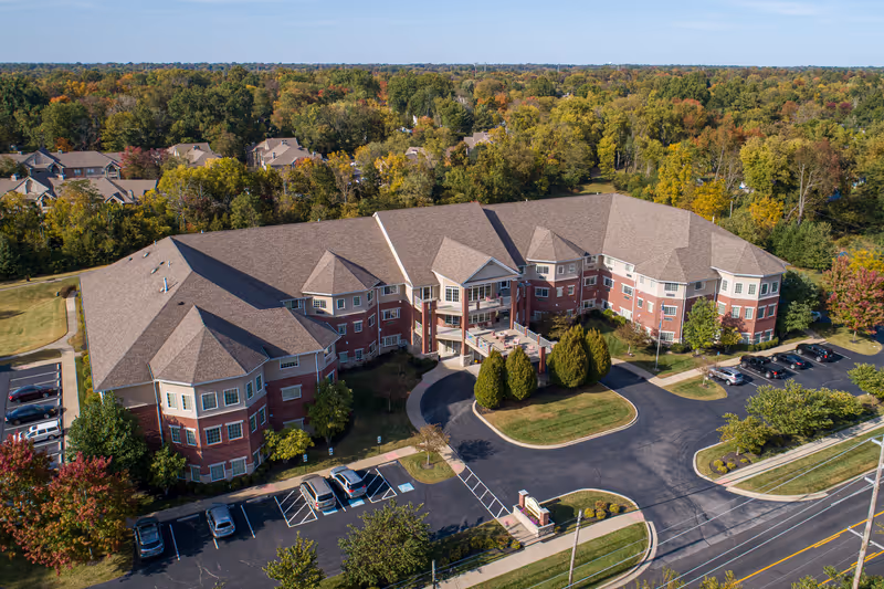 Aerial view of a three-story red-brick senior living building with a circular driveway, parking lots, and surrounding trees.