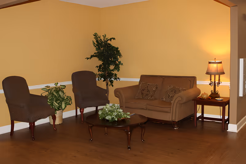 A cozy living room area with two brown armchairs, a brown loveseat with two decorative pillows, a round wooden coffee table with a floral arrangement, a side table with a lamp, and two potted plants against a yellow wall.
