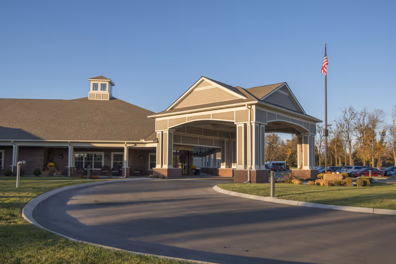 Exterior view of The Lantern at Morning Pointe Alzheimer's Center of Excellence in Danville, showing the building entrance with a covered driveway, an American flag on a flagpole, and a parking lot with cars in the background under a clear blue sky.
