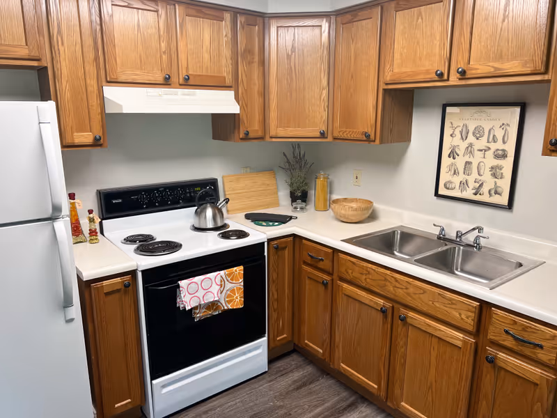 A kitchen with wooden cabinets, a white refrigerator, a white electric stove with a kettle on it, a double stainless steel sink, and various kitchen items including a cutting board, a bowl, a jar of pasta, and a framed vegetable garden print on the wall.