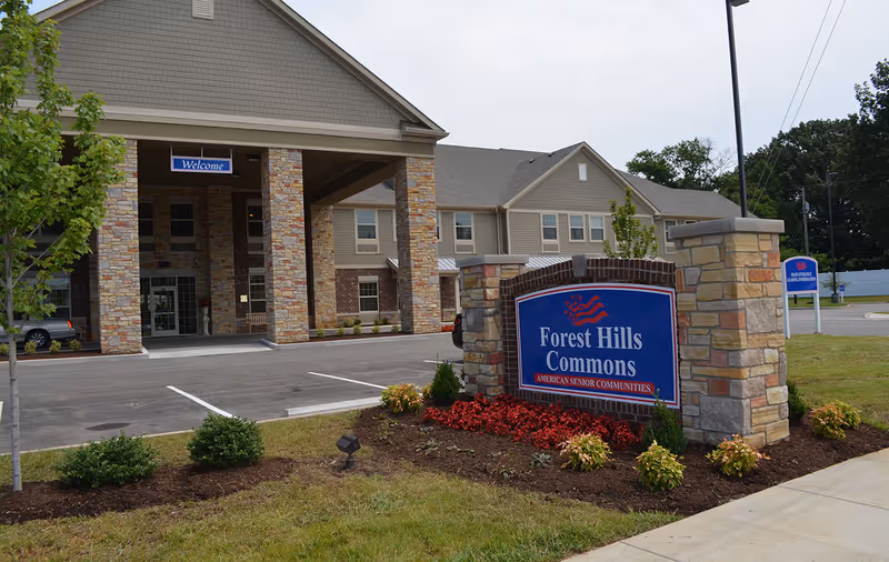 Exterior view of Forest Hills Commons senior living community showing the main entrance with stone pillars and a covered drop-off area. A large blue sign with red and white accents reads 'Forest Hills Commons American Senior Communities' surrounded by landscaping with red and green plants. The building is two stories with beige siding and brick accents.