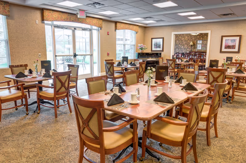 A bright and spacious dining room with multiple wooden tables and chairs arranged neatly. Each table is set with white cups, black napkins, silverware, and small flower vases. Large windows and glass doors allow natural light to fill the room, and framed artwork decorates the walls. The room has a warm and inviting atmosphere.