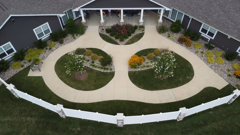 Aerial view of the entrance area of Cooper Trail Senior Living showing a curved concrete walkway with landscaped garden beds containing bushes and flowers, bordered by a white fence and green lawn, with the building facade in the background.
