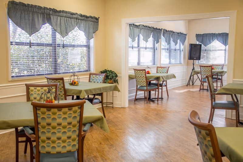 A bright dining area with multiple tables covered in green tablecloths and chairs with patterned upholstery. Large windows with blue valance curtains let in natural light, and there are small decorative centerpieces on the tables. The room has wood flooring and light-colored walls.