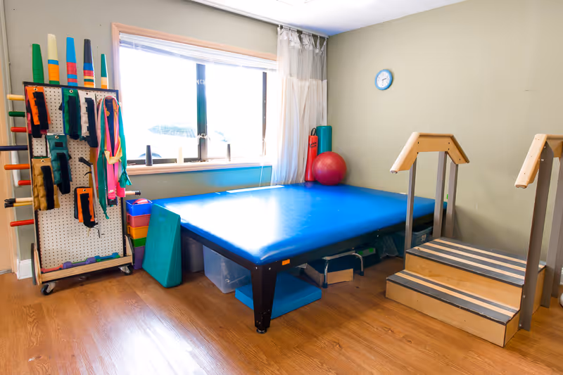 A therapy room with a large blue padded table, wooden steps with handrails, various exercise and therapy equipment including resistance bands, foam rollers, and a red exercise ball, all set against a light green wall with a clock and a window with white curtains.