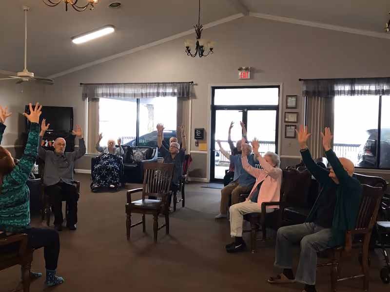 A group of elderly people seated in a spacious common room with high ceilings, raising their arms as if participating in a group exercise or activity. The room has large windows with sheer curtains, a TV on a stand, and wooden chairs arranged in a circle.