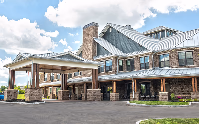 Exterior view of Dominion Senior Living of Florence, showing a large brick building with multiple windows, a metal roof, and a covered entrance supported by brick and wooden pillars. The driveway and landscaped areas with grass and flowers are visible under a partly cloudy sky.