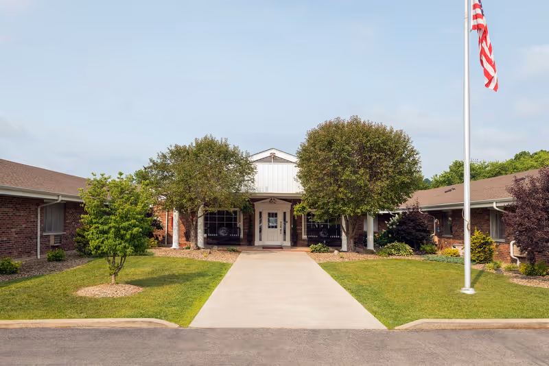Front exterior view of a single-story brick building with a central entrance flanked by two trees and a concrete walkway leading to the door. There is a flagpole with an American flag on the right side and well-maintained grass and shrubs surrounding the building.