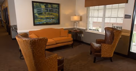 A cozy seating area in a senior living facility with an orange sofa, two brown patterned armchairs, a side table with a lamp, a large window with blinds, and a framed painting of a bridge over a stream on the wall.
