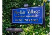 Blue sign that reads "Mayfair Village Retirement Community Front Entrance" set among green foliage.