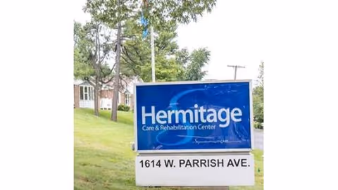 Outdoor view of a blue and white sign for Hermitage Care & Rehabilitation Center located at 1614 W. Parrish Ave, with trees and a building in the background.