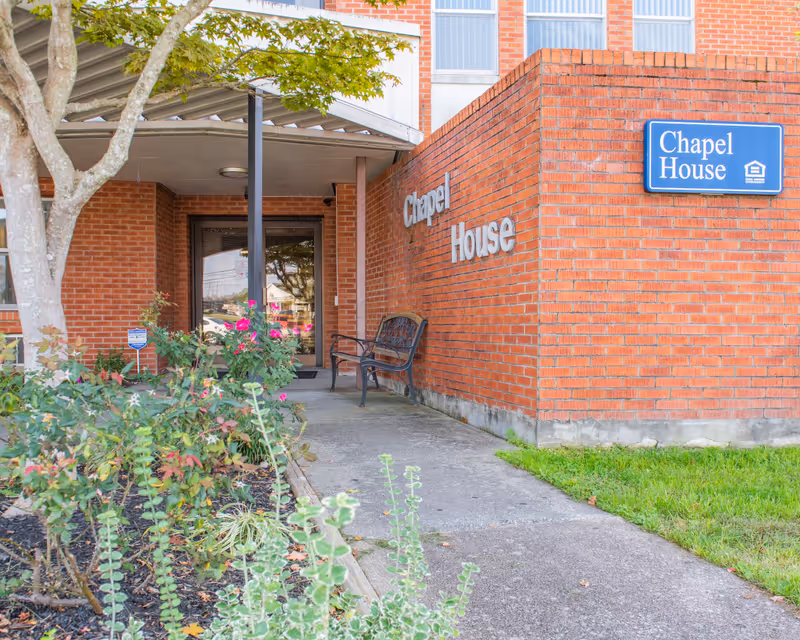 Entrance to Chapel House Corbin, a Christian Care Community, showing a brick building with a covered walkway, a bench, and some flowering plants near the entrance door. The building has a blue sign with white text reading 'Chapel House'.