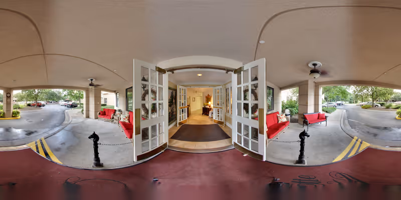 Covered entrance area of The Lafayette senior living facility with open double glass doors leading inside. There are red cushioned benches on both sides under the covered area, ceiling fans above, and a parking lot with cars and trees visible outside.