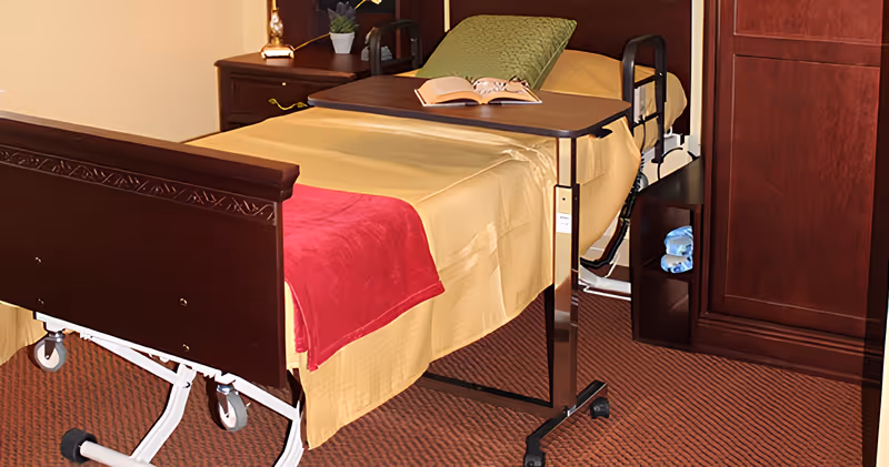 A hospital-style bed with wooden headboard and footboard in a furnished bedroom, with a yellow bedspread, red throw, overbed table holding an open book, nightstand and cabinet.