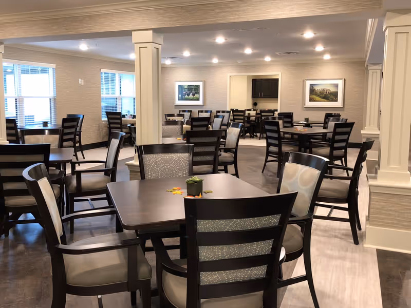 A spacious dining room with multiple dark wooden tables and chairs arranged neatly. The room has light-colored walls, large windows letting in natural light, framed pictures on the walls, and recessed ceiling lights. There are decorative columns and a small plant centerpiece on the closest table.
