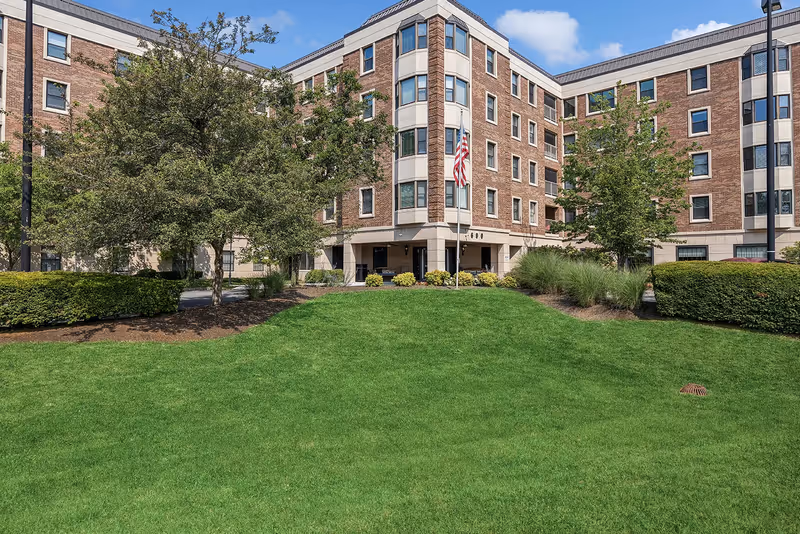 Front exterior of a multi-story brick senior living building with a green lawn, trees, and an American flag at the entrance.