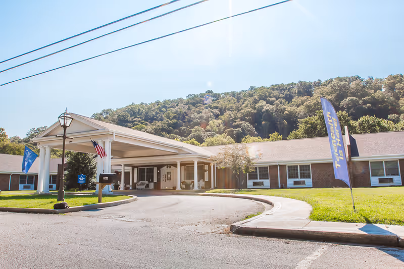 Exterior view of Lee County Care and Rehabilitation Center showing a single-story brick building with a covered entrance supported by white columns. There are flags near the entrance, including an American flag and a blue flag with text. The building is surrounded by green grass and trees, with a wooded hill in the background under a clear sky.