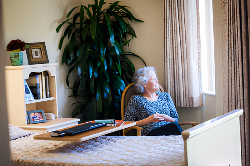 An elderly woman sits in a chair by a window in a nursing facility room with a bed, bedside table, and a large potted plant.