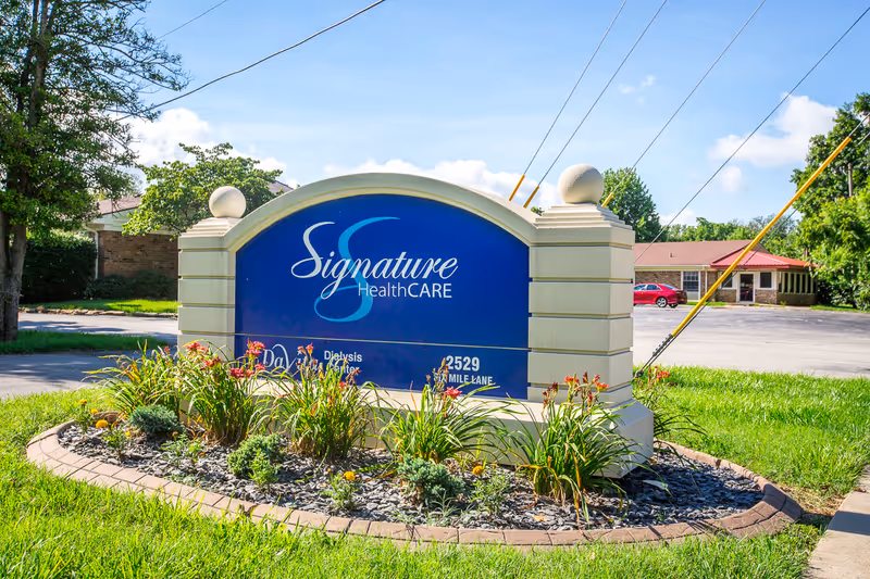 Outdoor view of a large blue and beige sign for Signature HealthCARE, surrounded by a landscaped area with flowers and greenery, with a building and parking lot in the background under a clear sky.