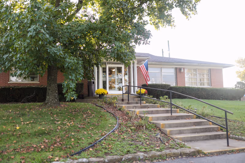 Exterior view of a single-story brick building with a front entrance featuring a glass door, flanked by two white columns. There is a concrete staircase with a black handrail leading up to the entrance, and an American flag is displayed near the door. The building is surrounded by green grass, a large tree, and some bushes.
