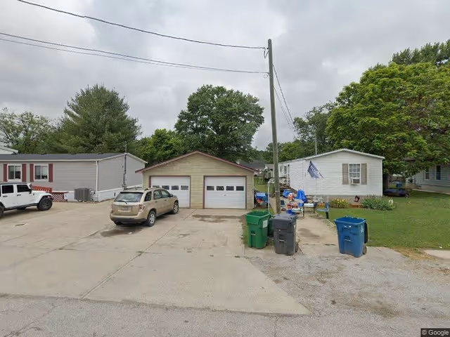 View of a residential area with a small garage with two white doors in the center, a beige car parked in front of it, and two single-story houses on either side. There are several trash bins and recycling containers near the garage, a utility pole with wires, and trees in the background under a cloudy sky.