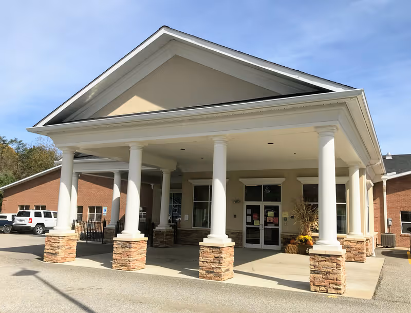 Front entrance of Carter Nursing and Rehabilitation facility featuring a covered drop-off area supported by white columns with stone bases, a beige building facade, and a brick side wall. There are some fall decorations near the entrance doors and a few parked vehicles visible on the left side.