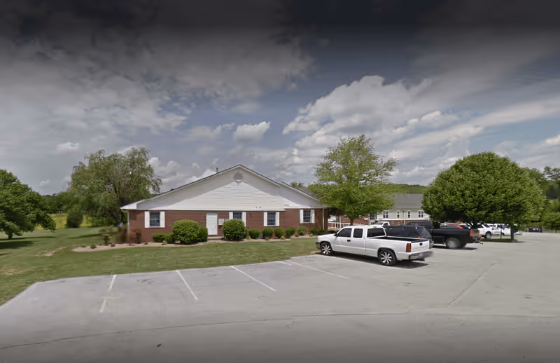 Exterior view of a single-story brick and siding building with a parking lot in front. Several vehicles are parked, and there are green trees and grass surrounding the building under a partly cloudy sky.