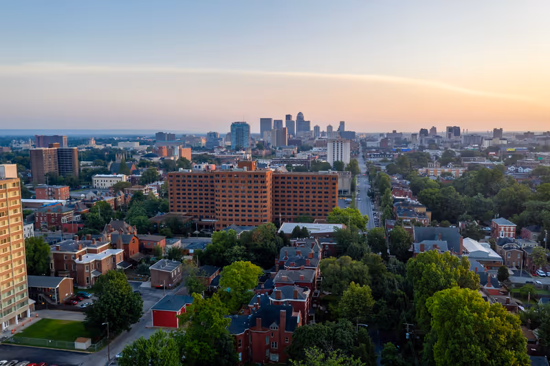 Aerial view of an urban area at sunset showing a mix of residential buildings, trees, and a large brick apartment complex in the foreground with a city skyline in the distance under a clear sky.