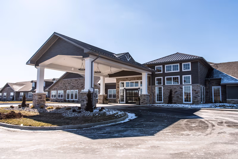 Front exterior of a senior living facility with a covered porte-cochere, stone and siding facade, and light snow on the ground.