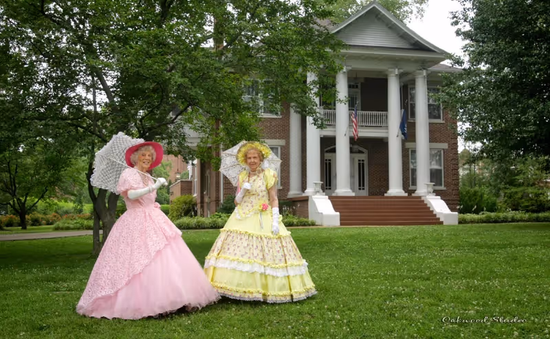 Two women in Victorian-style dresses holding parasols stand on the lawn in front of a large columned brick building.
