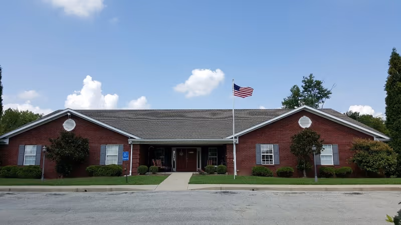 Front exterior view of a single-story red brick building with a gray shingled roof, an American flag on a flagpole in front, and a paved driveway leading to the entrance. The building is surrounded by green bushes and trees under a blue sky with some clouds.