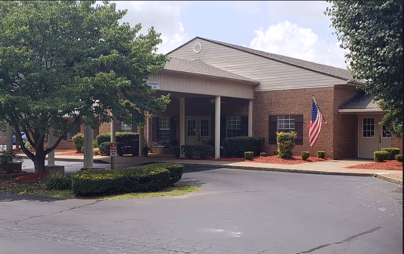 Exterior view of a single-story brick building with a covered entrance supported by white columns. There are neatly trimmed bushes and trees around the building, an American flag mounted on the wall, and a paved driveway in front.