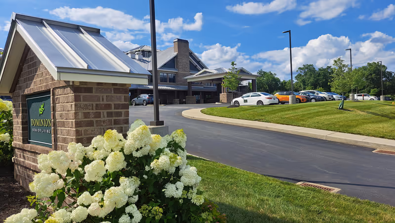 Exterior view of Dominion Senior Living facility with brick sign, manicured lawn, and parked cars.