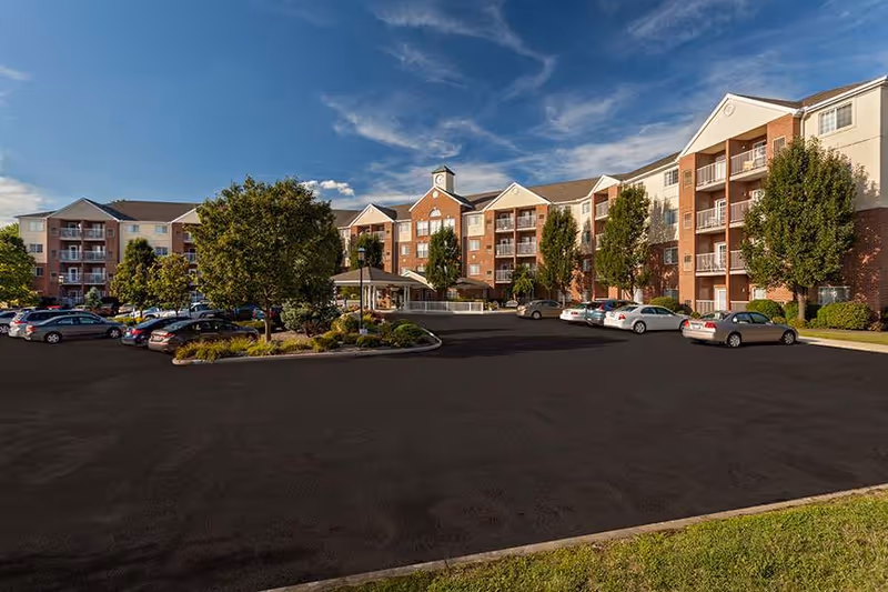 Exterior view of a multi-story senior living facility with brick and beige siding, balconies, and a parking lot with several cars. There are trees and landscaped areas around the building under a partly cloudy blue sky.