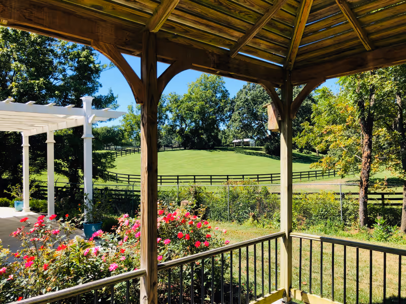 View from a wooden gazebo overlooking a garden with blooming pink and red flowers, a white pergola, green grassy fields, trees, and a wooden fence under a clear blue sky.