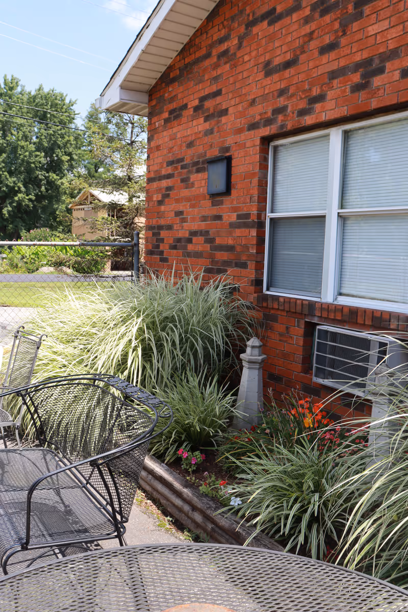 Outdoor patio area next to a red brick building with a window air conditioning unit. The patio has black metal mesh tables and chairs. There are ornamental grasses and flowering plants along the building's foundation, with a small decorative stone pillar among the plants. A chain-link fence and trees are visible in the background under a clear sky.