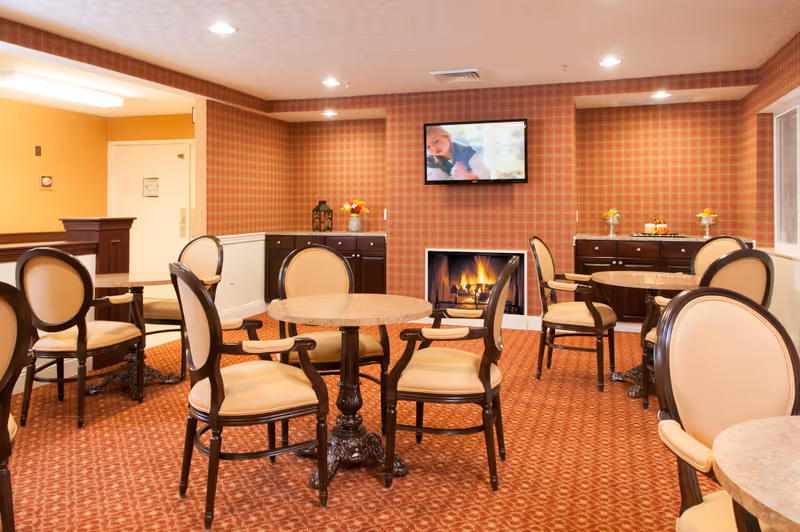Warm communal lounge with circular tables, upholstered chairs, fireplace, and wall-mounted TV.