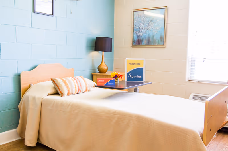 A single bedroom in a senior facility with a bed, nightstand, lamp, and window.