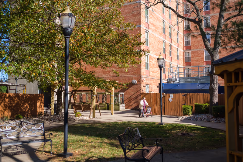 Outdoor courtyard area at Friendship House Louisville with benches, lamp posts, trees, and a person walking with a walker near a brick building.