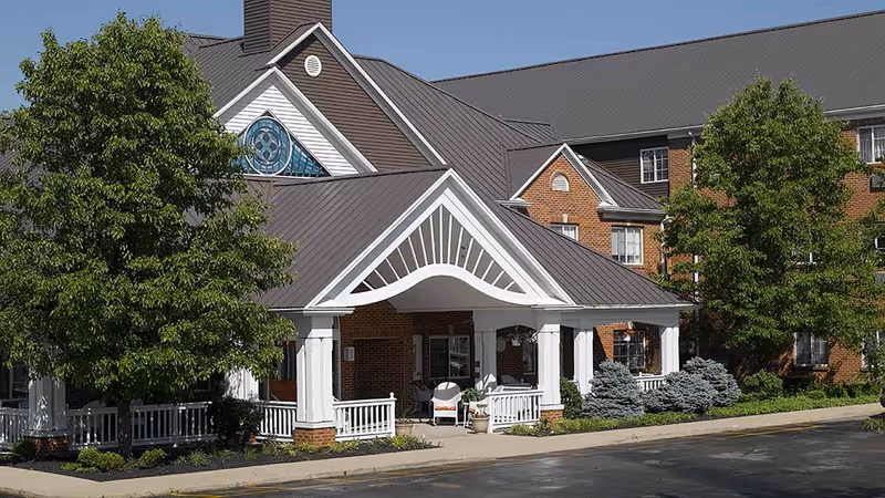 Front entrance of a brick senior living building with a white-columned covered portico, trees, and landscaping.