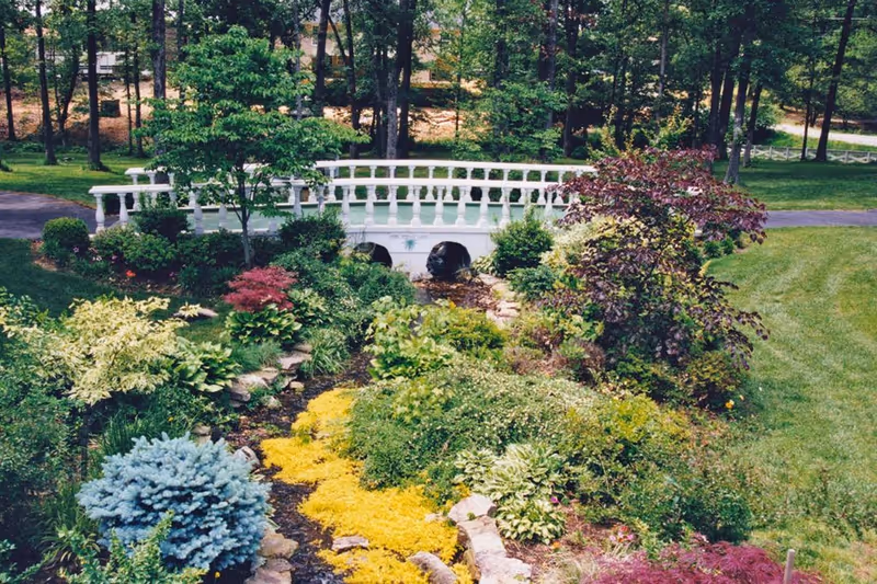 A lush garden area with various green shrubs, colorful plants, and a small white decorative bridge over a dry creek bed, surrounded by trees and a grassy lawn.