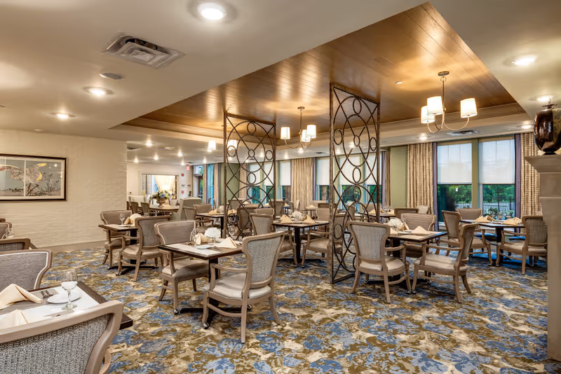 A spacious dining room with multiple tables and chairs arranged neatly. Each table is set with napkins, plates, and glasses. The room features a patterned carpet in blue and brown tones, decorative metal room dividers, large windows with curtains, and warm ceiling lighting fixtures.